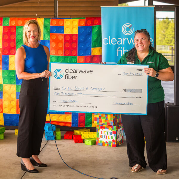 Two people hold a $1,000 check from Clearwave Fiber to Girl Scouts of Gateway, standing in front of colorful LEGO-like blocks.